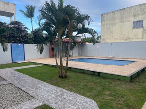 A swimming pool with a tiled deck, a grassy area, and a building with a blue door. - Residencial Brasil Coroa (Santa Cruz Cabrália)