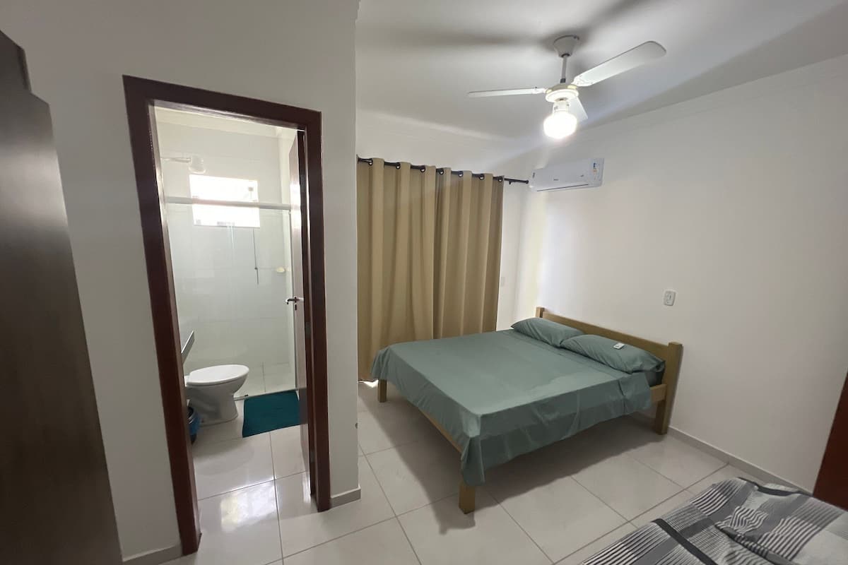 Un dormitorio con cama, ventilador de techo, ventana con cortinas y baño visible a través de una puerta abierta.