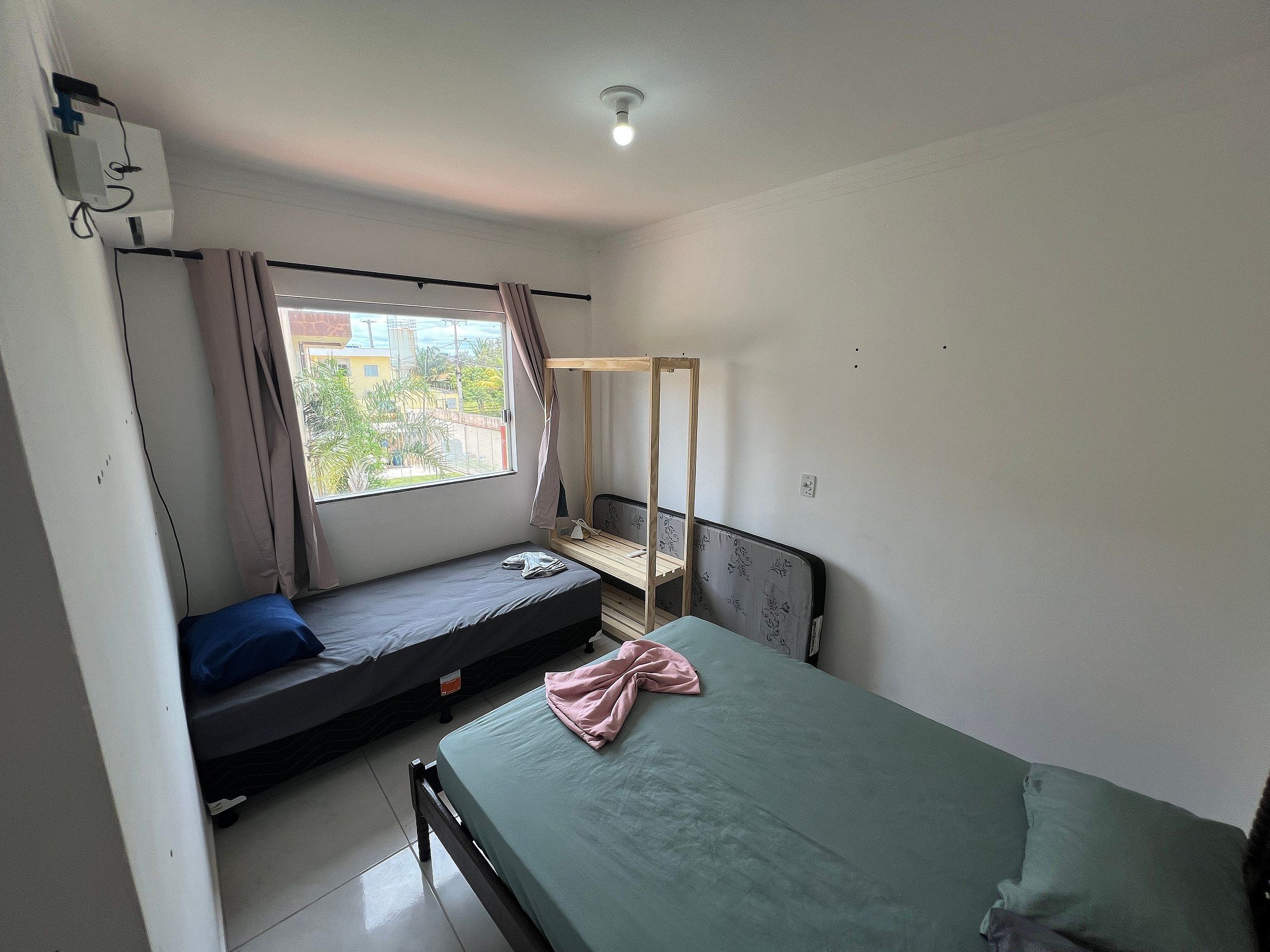 A dormitory room with two beds, a window with curtains, a ceiling light, and a power outlet on the wall.