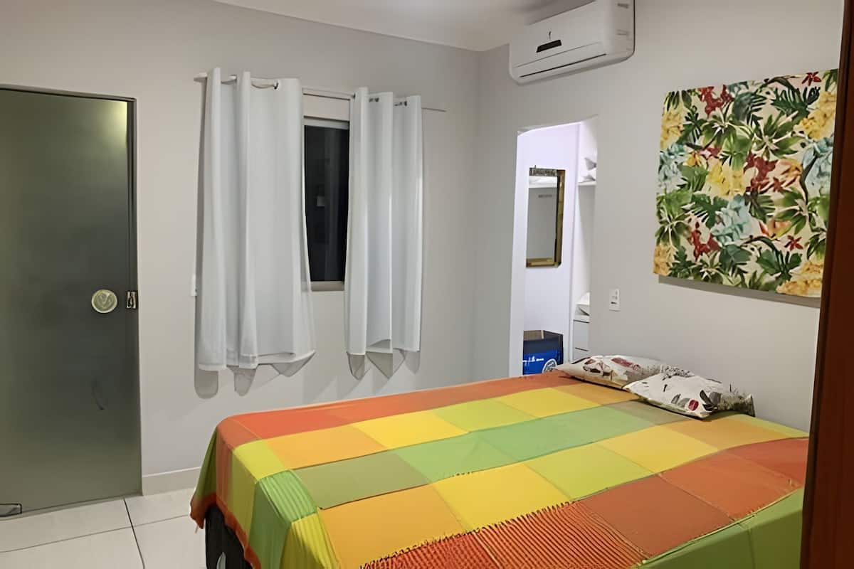 Un dormitorio con cama, puerta, ventana con cortinas, equipo de aire acondicionado y un cuadro floral en la pared.
