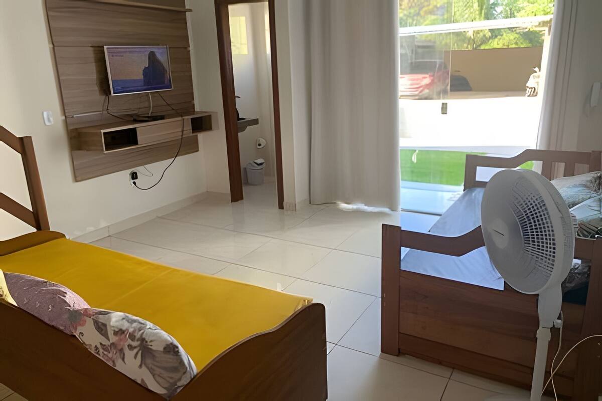 Una habitación con cama, ventilador, televisor sobre una estantería de madera y vistas al exterior.