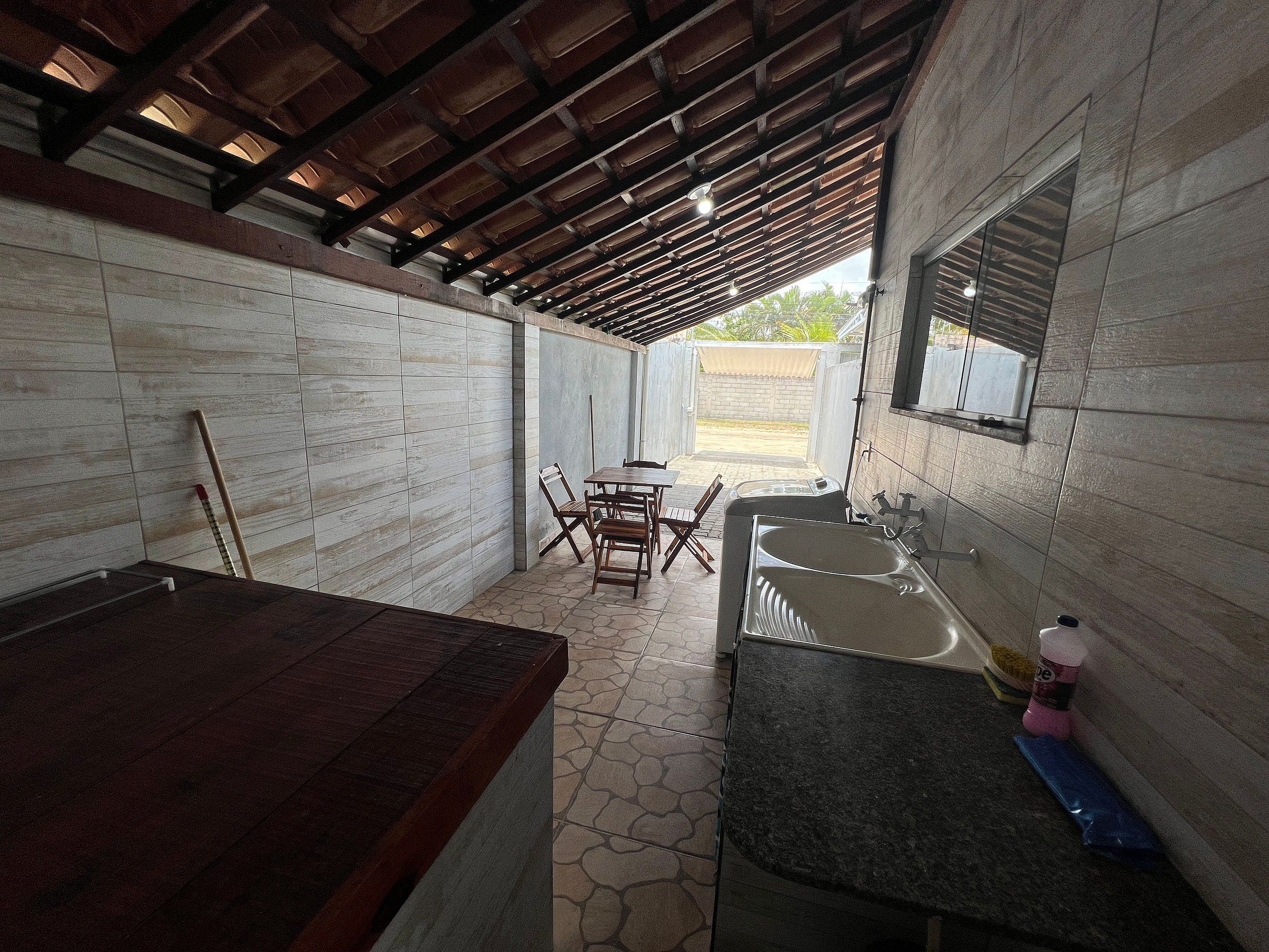 Uma área de cozinha ao ar livre coberta, com uma mesa de jantar e cadeiras, pia e uma mesa de madeira.