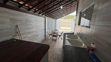 Uma área de cozinha ao ar livre coberta, com uma mesa de jantar e cadeiras, pia e uma mesa de madeira.