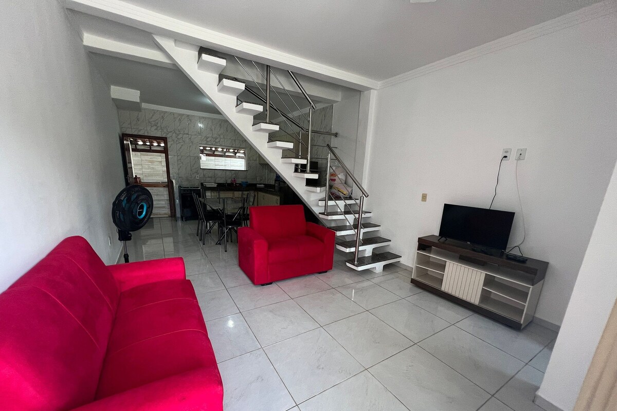 Ein Wohnzimmer mit einem roten Sofa, einem Fernseher auf einem Holzständer, einer Treppe und einem Essbereich mit Stühlen und einem Tisch.