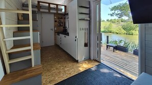 A modern interior with a wooden staircase, a kitchen with white cabinets, and a view of a lake through a glass door. - Tiny House mit Terrasse, Grill & Kamin Direkt am Naturteich - Best Price! (Grossharthau)