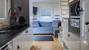 A compact kitchen with a stove, sink, and cabinets, a small seating area with a sofa and pillows, and a loft bed with a ladder. - Tiny House mit Terrasse, Grill & Kamin Direkt am Naturteich - Best Price! (Grossharthau)
