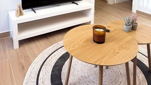 Moderni olohuone, jossa on valkoinen TV-taso ja sen päällä litteänäyttöinen televisio, pyöreä puinen sohvapöytä, jonka päällä on kynttilä, sekä pieni ruukkukasvi.
