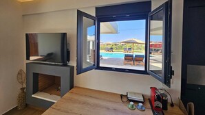 A modern room with a TV, a fireplace, a coffee maker, and a view of a pool and outdoor seating. - Mat Suites (rhodes)