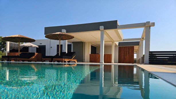 A clear swimming pool with lounge chairs and umbrellas, surrounded by a modern building with white walls and wooden accents. - Mat Suites (rhodes)