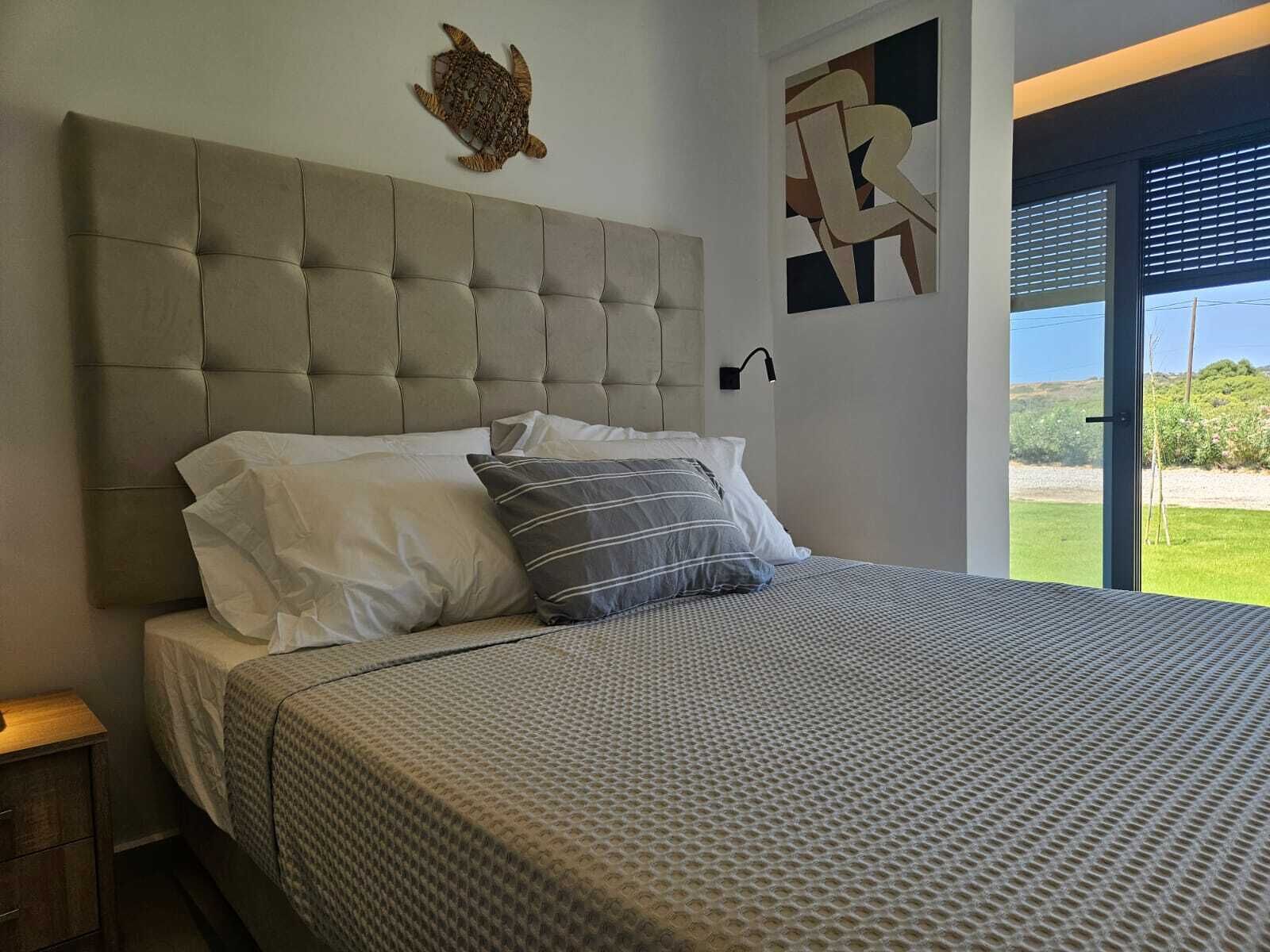 Een slaapkamer met een gestuufd hoofdeinde, een bed met beddengoed met een patroon, een wanddecoratie van een schildpad, en een raam met uitzicht op groen.