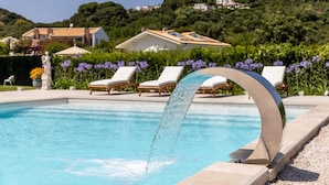 A swimming pool with a water feature, lounge chairs, and a view of houses and greenery in the background.