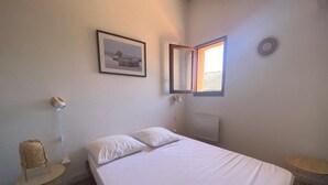 Ein Schlafzimmer mit einem Bett, einer Nachttischlampe, einem runden Tisch, einem Stuhl, einem gerahmten Bild, einem Fenster und einer Heizung.