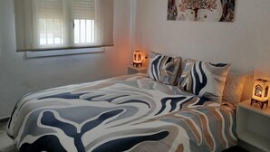 Een slaapkamer met een bed, twee bedlampjes, een raam met zonwering en een aan de muur hangend schilderij.
