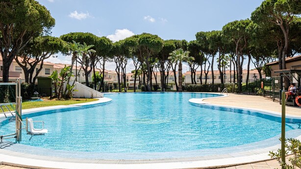 Une piscine entourée d’une terrasse, d’arbres et d’un bâtiment en arrière-plan.