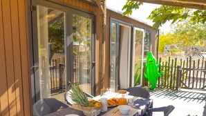 An outdoor dining area with a table set for two, surrounded by chairs, and a wooden deck with a glass door leading to a garden. - Mobile home with AC - Pets OK - 6 guests (Frontignan)