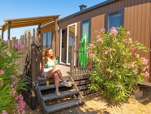 A wooden deck with a person sitting, a green gate, and pink flowering plants. - Mobile home for 8 guests, pets welcome (Frontignan)