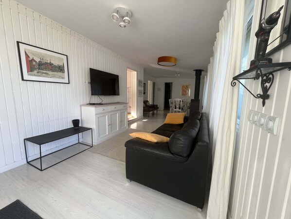 Un salon moderne avec un canapé en cuir noir, un téléviseur à écran plat, un meuble en bois et, au fond, un coin repas.