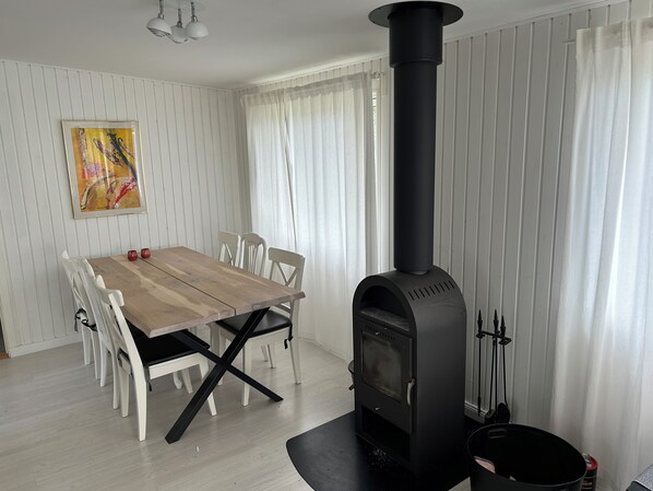 A dining area with a wooden table, white chairs, a black wood stove, and a painting on the wall. - Dejligt hus på Enø i Karrebæksminde (Karrebæksminde)