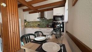 A dining area with a wooden table set for two, a kitchen in the background, and a wooden ceiling. - Charming house near the beach - 4 guests (Saint-Georges-d'Oléron)