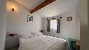 A bedroom with a bed, a window with curtains, a bedside table with a lamp, and a wooden beam ceiling. - Charming house near the beach - 4 guests (Saint-Georges-d'Oléron)