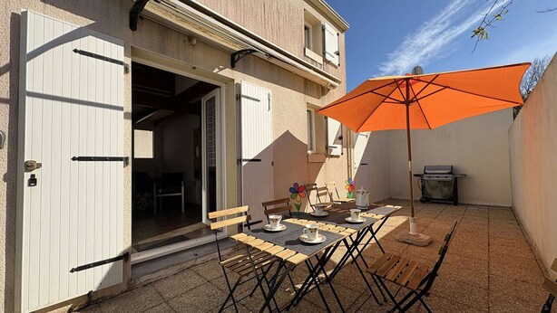 An outdoor patio with a table set for dining, a grill, and an open door leading to a room. - Charming house near the beach - 4 guests (Saint-Georges-d'Oléron)