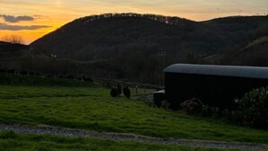 Paisaje rural con un campo cubierto de hierba, un edificio de techo oscuro y una colina al fondo.