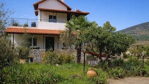 Uma casa de dois andares com telhado de telhas, rodeada por vegetação exuberante e uma paisagem acidentada.
