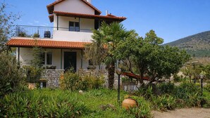 Una casa de dos pisos con techo de tejas, rodeada de exuberante vegetación y un paisaje montañoso.