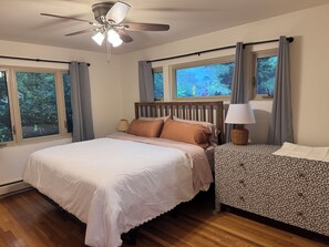 Ein Schlafzimmer mit einem Bett, einem Nachttisch, einer Lampe, einem Deckenventilator und großen Fenstern mit Vorhängen.