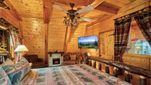 L’intérieur d’une cabane en rondins, avec une cheminée, un lit recouvert d’une literie à motifs, un téléviseur et un ventilateur de plafond.