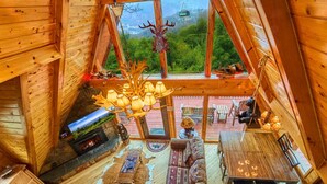 Une cabane en bois avec une chambre à coucher située en mezzanine, une cheminée, un coin repas et une vue sur l’extérieur.