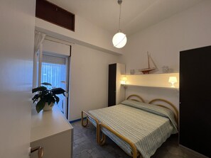 A bedroom with a bed, bedside lamps, a small shelf with decorative items, a potted plant, and a window with curtains. - Cozy Accommodation Just 250mt From the Beach (Lignano Sabbiadoro)