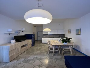 A modern kitchen with white cabinets, a dining table with chairs, a TV, and a large pendant light. - Cozy Accommodation Just 250mt From the Beach (Lignano Sabbiadoro)