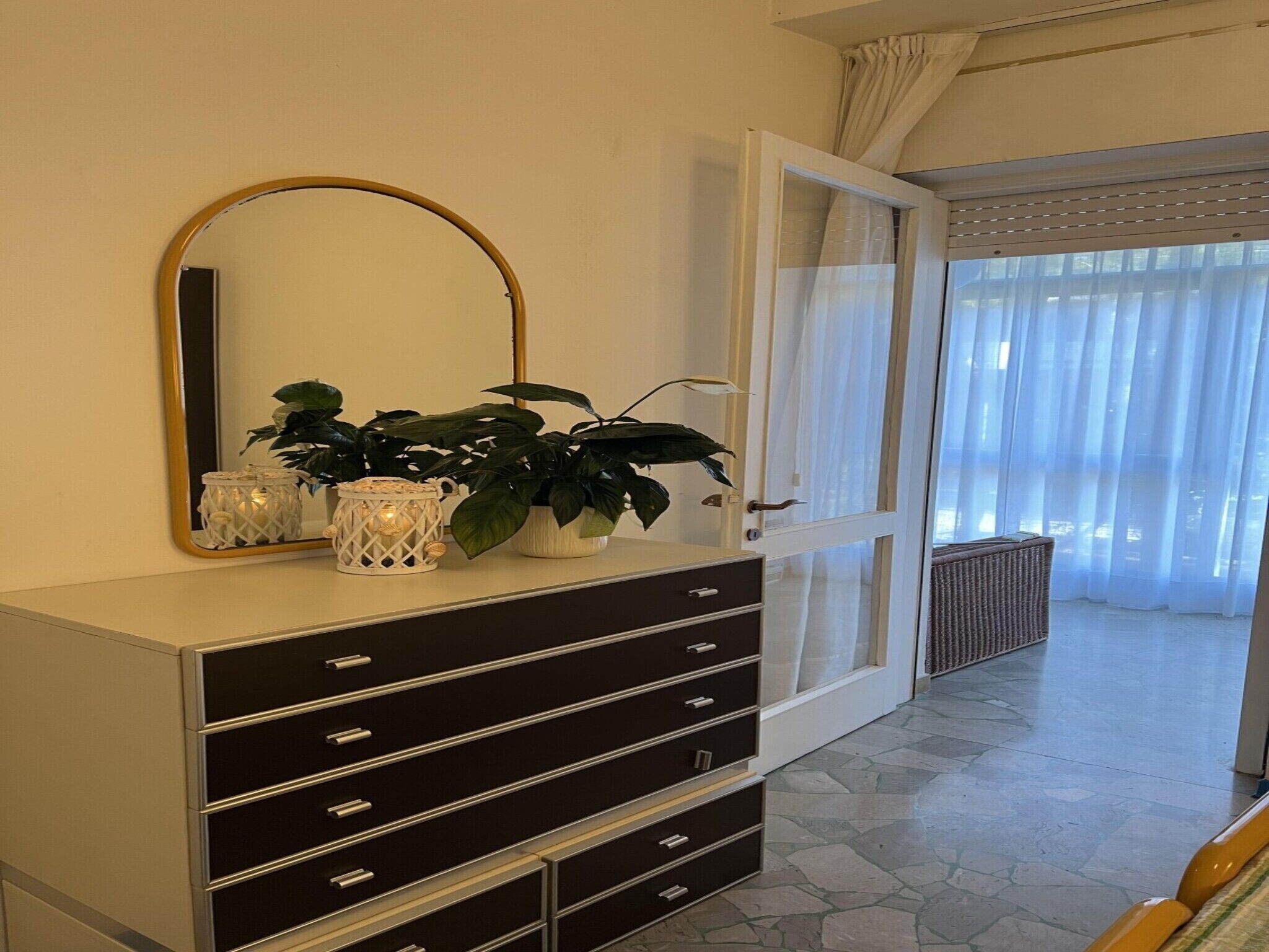 A room with a large mirror, a dresser with drawers, a potted plant, and a view of a balcony with curtains.