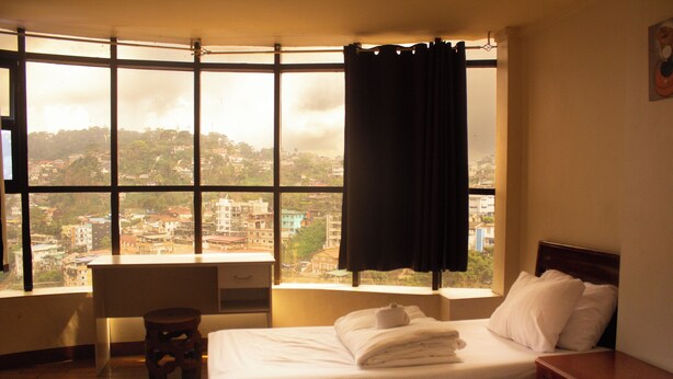 A hotel room with a large window, a bed, a bedside table, and a small stool.