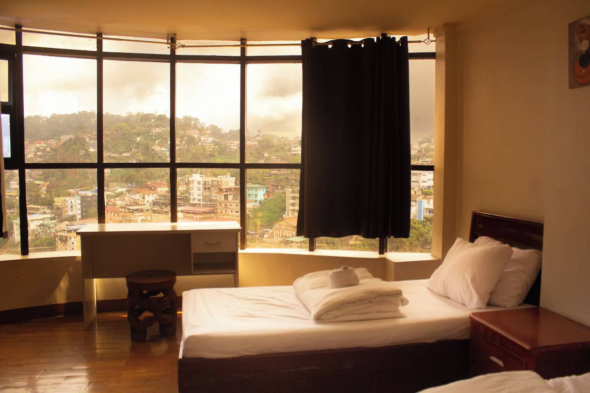 Una habitación de hotel con un ventanal grande, una cama, una mesita de noche y un pequeño taburete.
