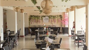 A restaurant with a central wall decorated with greenery and hanging lights, tables and chairs arranged for dining, and a sign indicating the restaurant name.