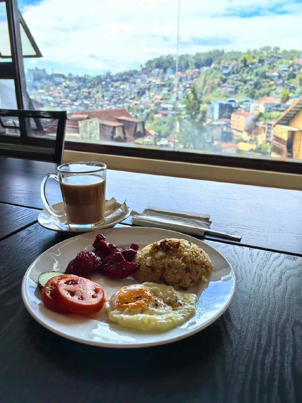 Une assiette de nourriture et une tasse de café sont posées sur une table près d’une fenêtre donnant sur un paysage urbain.