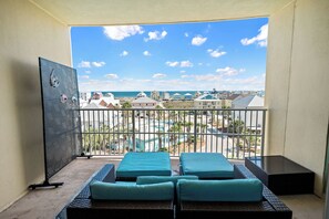 A balcony with four blue cushioned chairs, a metal gate, and a view of a beach and buildings. - Indies 406 | 1BR/1BA Gulf-View Condo | Sleeps 4 | Balcony & Pool Access (GULF SHORES)