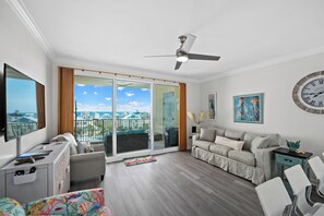 A modern living room with a large window, a ceiling fan, a sofa, a coffee table, and a TV. - Indies 406 | 1BR/1BA Gulf-View Condo | Sleeps 4 | Balcony & Pool Access (GULF SHORES)