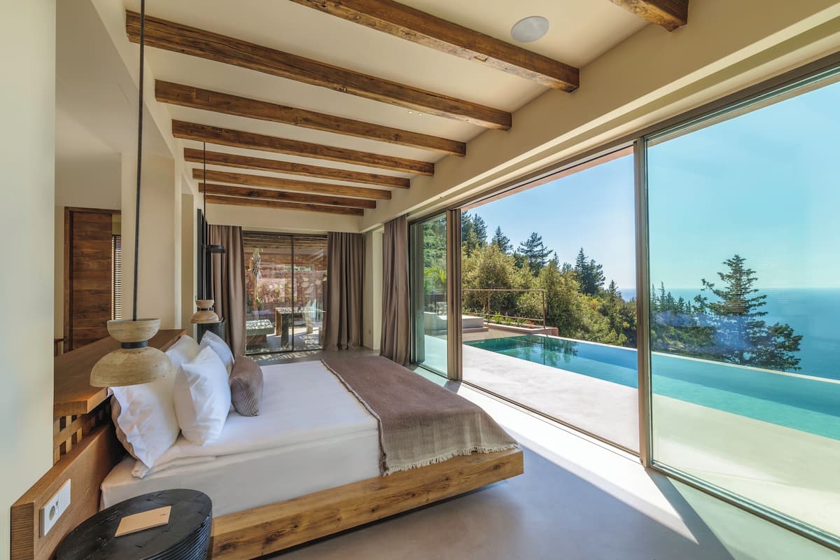 A bedroom with a large window, wooden beams, a bed, and a view of a swimming pool and trees.