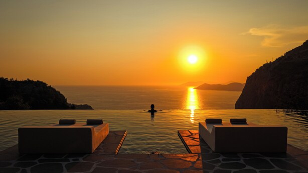 A sunset view from an infinity pool with lounge chairs and a person in the water. - Klay Hotel Faralya Adults Only (Fethiye)