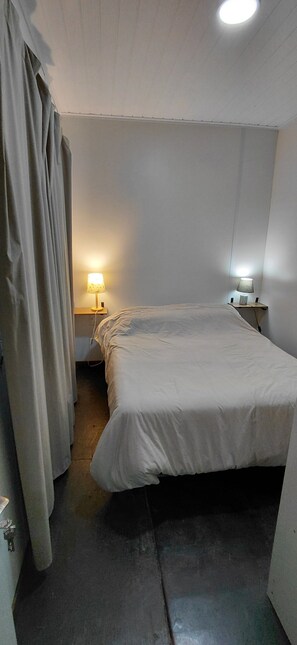 A small, dimly lit room with a single bed, a nightstand with a lamp, and a curtain partially drawn. - Modern Eco-Container Retreat: 2 Blocks from the Ocean, 1-bedroom house with AC. (Ba Buenos Aires ,Maldonado)