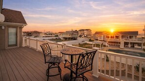 A wooden deck with two chairs and a table, overlooking a residential area with houses and a swimming pool during sunset. - New / Oceanview / Heated Pool • Steps to Beach (Virginia Beach)