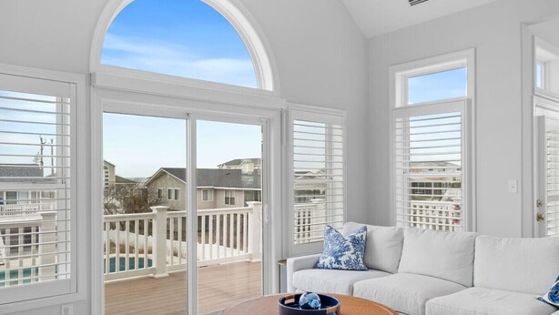 A spacious room with a large sliding glass door leading to a balcony, a white sofa with blue pillows, and a round wooden table with a bowl and a glass. - New / Oceanview / Heated Pool • Steps to Beach (Virginia Beach)