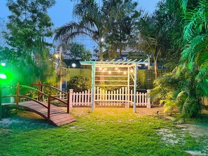 A garden with a wooden bridge, a white picket fence, and a pergola with string lights.