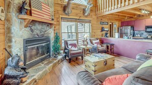A log cabin interior with a stone fireplace, wooden walls, a bear statue, a bear-themed chair, a wooden chest, and a kitchen area. - Spacious Log Cabin w/ Stunning Views in Blue Ridge (Blue Ridge)