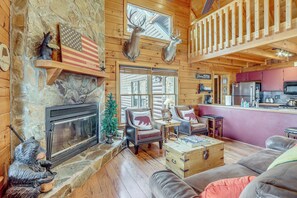 A log cabin interior with a stone fireplace, wooden walls, a bear statue, a bear-themed chair, a wooden chest, and a kitchen area. - Spacious Log Cabin w/ Stunning Views in Blue Ridge (Blue Ridge)
