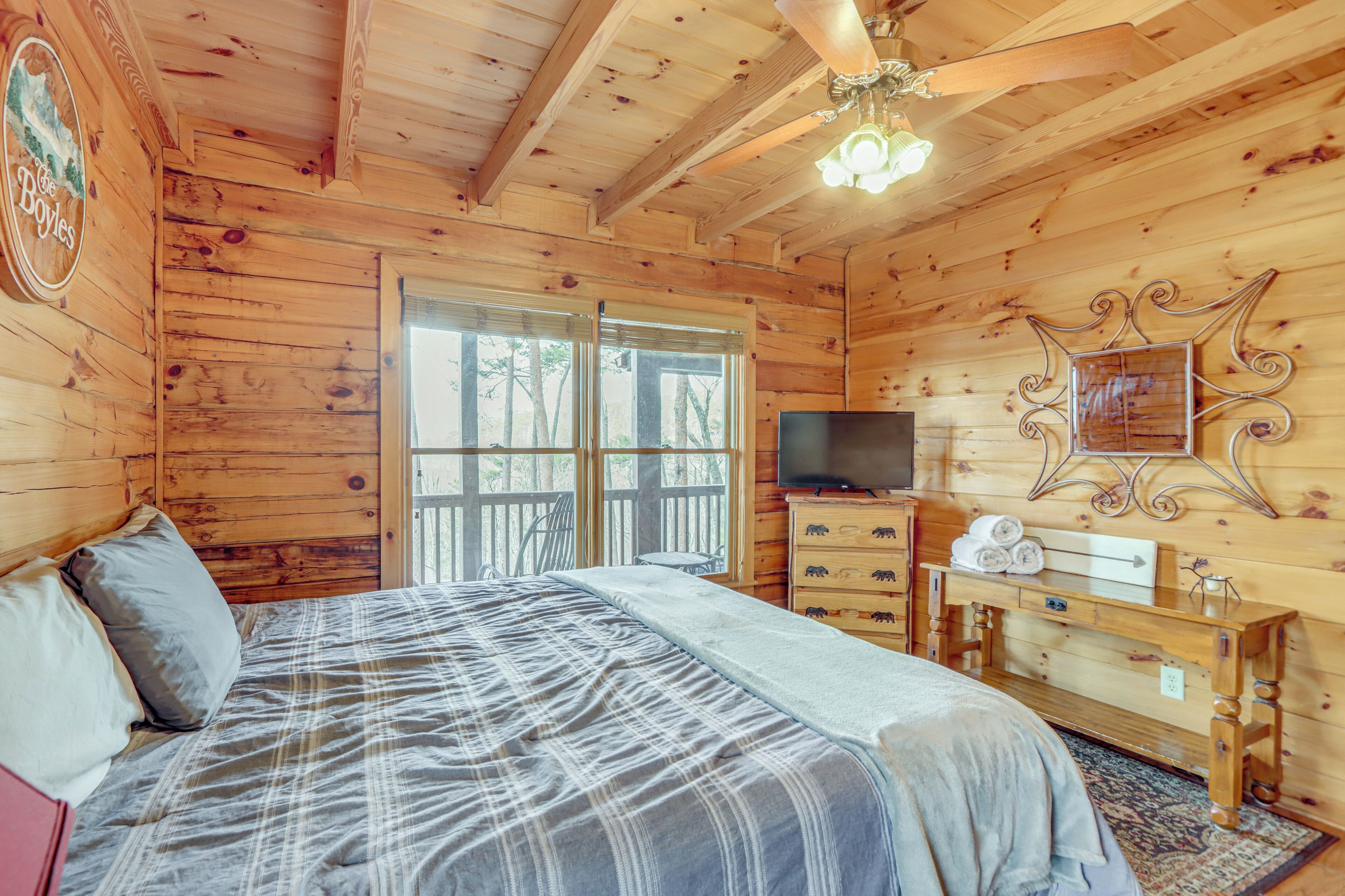 A wooden cabin bedroom with a bed, dresser, TV, and a balcony with a view of trees.