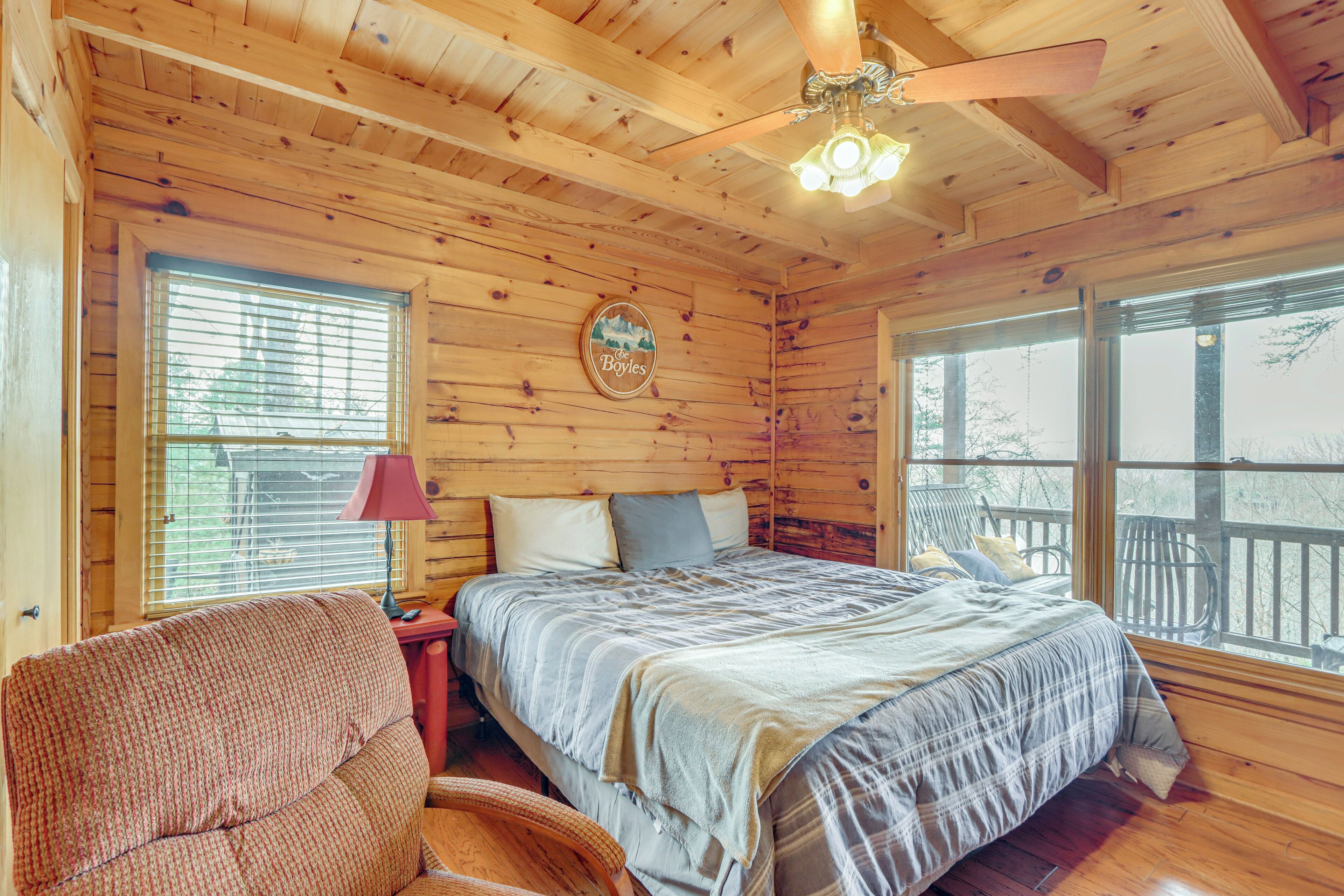 A wooden cabin bedroom with a bed, a chair, a nightstand, a ceiling fan, and a window with a view.